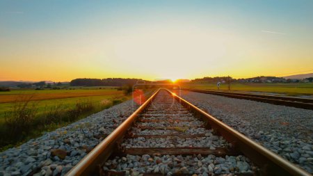 Téléchargez les photos : LOW ANGLE: Beautiful orange glowing railroad tracks in golden autumn sunlight. Countryside railway passing picturesque hilly landscape. Detailed view of railway tracks and beams for train transport. - en image libre de droit