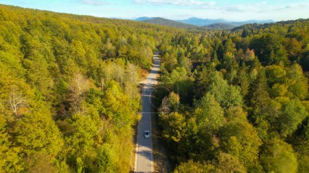 Téléchargez les photos : Country road winding through hilly forested landscape and a driving car. Picturesque countryside and enjoyable travel through lush autumn woodland in beautiful shades of early fall season. - en image libre de droit
