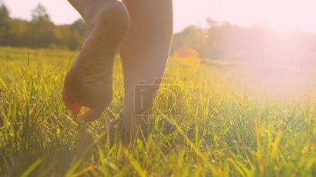 Foto de CERRAR, ÁNGULO BAJO: Vista trasera de la señora paseando la hierba descalza en luz dorada. Una joven descalza pisando un prado verde. Momento despreocupado y relajante en la naturaleza en una hermosa mañana soleada. - Imagen libre de derechos