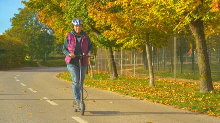 Foto de Señora mayor disfrutando de montar a caballo con scooter eléctrico en un hermoso día de otoño. Mujer de edad avanzada que utiliza el medio de transporte ecológico extremo moderno. Precioso día de otoño para pasear con e-scooter por la ciudad. - Imagen libre de derechos