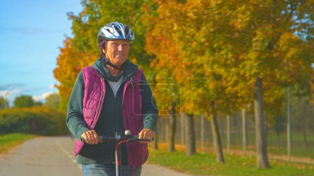 Foto de Cheerful senior woman enjoying driving with e-scooter on an autumn day. Elderly lady riding electric scooter on a cycle path along autumn tree-lined avenue. Lovely fall day for scooter ride. - Imagen libre de derechos