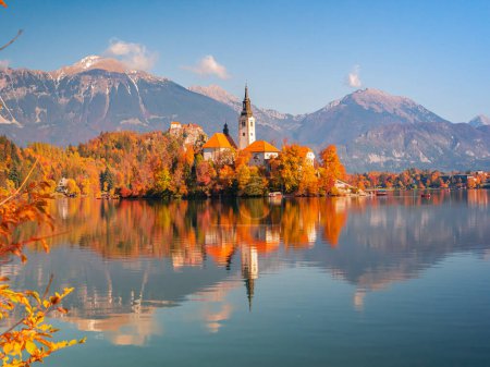 Téléchargez les photos : Beautiful church on a small island in the middle of lake Bled in autumn colors. World renowned travel destination in beautiful colors of fall season. Picturesque view of mountain lake with island. - en image libre de droit