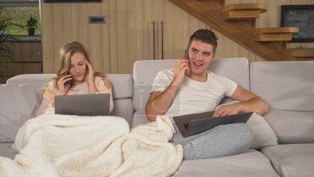 Téléchargez les photos : Couple using laptops and smartphones for remote work in home isolation. Married couple making business calls and other office work from their home couch office while being in quarantine. - en image libre de droit