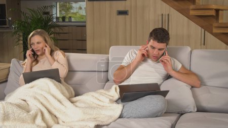 Foto de Couple disturbing each other while having business meeting phone calls. Married partners annoying and distracting each other at remote work during isolation. Young couple trying work at home - Imagen libre de derechos
