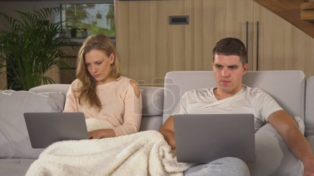 Photo for Student couple in quarantine trying to do their study responsibilities. Young couple siting on couch covered with blanket and using laptops to fulfil their study obligations in isolation. - Royalty Free Image