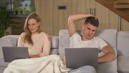 Foto de Couple using laptops for remote work at home living room. Young man and woman siting on couch covered with blanket and using laptops to fulfil work responsibilities during isolation - Imagen libre de derechos