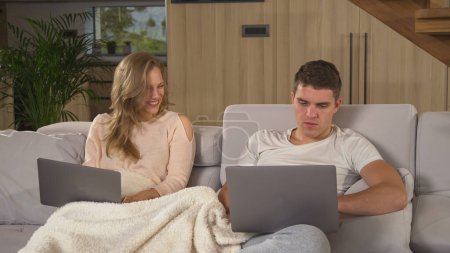 Foto de Two students in isolation using laptops for remote study at home. Young man and woman siting on couch covered with blanket and using laptops to fulfil study responsibilities during quarantine - Imagen libre de derechos