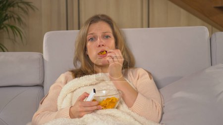 Téléchargez les photos : Pretty woman becoming stressed at watching unpleasant TV news report. Pretty woman covered with blanket and eating snacks while following TV news. Woman on a comfy sofa watching TV report. - en image libre de droit