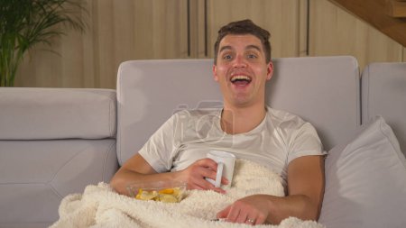 Photo for Man enjoying on a comfy couch and laughing while watching comedy movie. Man eating snacks, drinking tea and smiling while watching humorous entertainment show on TV on a long winter evening. - Royalty Free Image