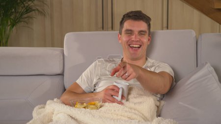 Photo for Handsome guy laughing and smiling while watching entertaining TV show. Man is enjoying on a comfy couch, eating snacks and watching humorous comedy movie on television on a winter evening. - Royalty Free Image