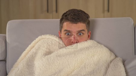 Photo for Young man watching horror movie and hides under blanket at scary scene. Young guy getting scared while watching TV movie. Male person chilling in front of television on a winter evening. - Royalty Free Image
