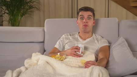 Téléchargez les photos : Man with surprised look when seeing shocking news on television report. Handsome guy covered with warm blanket getting upset while following TV news. Man on a comfy sofa watching TV report. - en image libre de droit