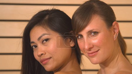 Photo for Two young beautiful Caucasian and Asian women posing for the camera. Attractive females standing side-by-side looking into camera. Girlfriends having relaxing beauty treatment in wellness. - Royalty Free Image