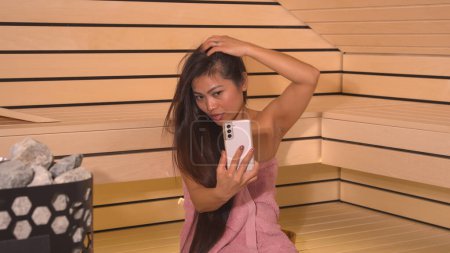 Téléchargez les photos : Good-looking Asian woman taking selfie while having a sauna treatment. Pretty lady sitting in Finnish sauna and using smartphone for photographing herself. Carefree weekend at beauty centre. - en image libre de droit