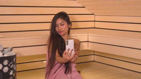 Téléchargez les photos : Beautiful Philippine woman taking selfies for social media in sauna. Pretty lady sitting in Finnish sauna and using smartphone for photographing herself. Beauty treatment at spa centre. - en image libre de droit