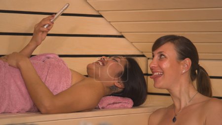 Foto de Smiling young women creating memories from relaxing at sauna and spa. Beautiful girlfriends wrapped in pink towels enjoying and having fun at wellness treatment in wooden Finnish sauna. - Imagen libre de derechos