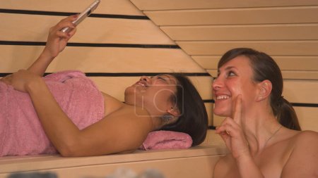 Téléchargez les photos : CLOSE UP: Cheerful Asian and Caucasian woman making a selfie in Finnish sauna. Beautiful girlfriends wrapped in pink towels enjoying and having fun at wellness spa treatment in wooden Finnish sauna. - en image libre de droit