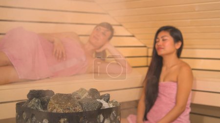 Foto de Steaming from sauna stones and beautiful ladies relaxing in background. Two attractive girlfriends wrapped in pink towels at wellness treatment, detoxifying the body in hot Finnish sauna. - Imagen libre de derechos