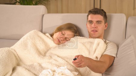 Foto de Young couple resting under blanket and watching TV while catching flu. Twosome having a seasonal cold and snuggling on cosy sofa. Man watching a movie while woman is being tired and sleeping - Imagen libre de derechos