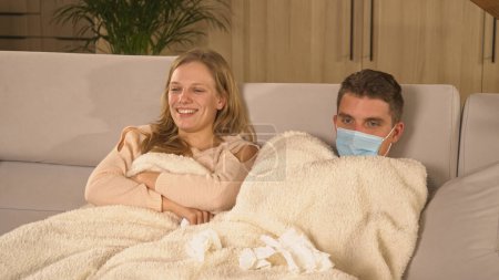 Foto de Lovely couple sitting on comfy sofa and getting over a seasonal cold. Winter colds and flu spreading around. Young guy and lady being on sick leave, covered with blanket and watching TV. - Imagen libre de derechos