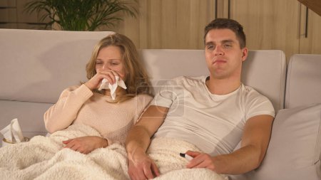 Photo for Woman with runny nose watching movie with partner during sick leave. Young couple getting over seasonal flu while lying on cosy couch under blanket. Autumn flu and colds spreading around. - Royalty Free Image