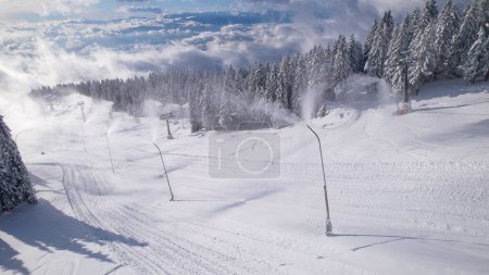 Téléchargez les photos : Snowmakers spraying artificial snow at ski resort with gorgeous view. Snow preparation for consolidating snow base at ski slopes and a long winter season. Winter wonderland in the mountains. - en image libre de droit