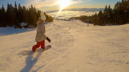 Téléchargez les photos : Woman with snowboard riding down the slope on a sunny winter day in mountains. Perfect conditions for snowboarding and skiing at snowy alpine ski resort with magnificent views above misty valley. - en image libre de droit