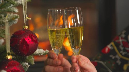 Foto de Festive champagne toast by the Christmas tree and fireplace. Idyllic home environment for celebrating Christmas Eve. Two hands toasting with champagne glasses on festive Evening. - Imagen libre de derechos