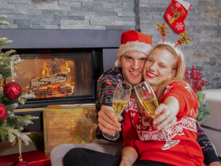 Téléchargez les photos : Happy young couple toasts with glasses of champagne on Christmas Eve. Cheerful man and woman enjoying and celebrating with champagne sitting in front of a fireplace by the Christmas tree. - en image libre de droit