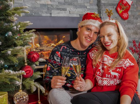 Photo for Smiling young couple toasts with glasses of champagne on New Years Eve. Happy young man and woman enjoying and celebrating with champagne sitting by Christmas tree in front of a fireplace. - Royalty Free Image