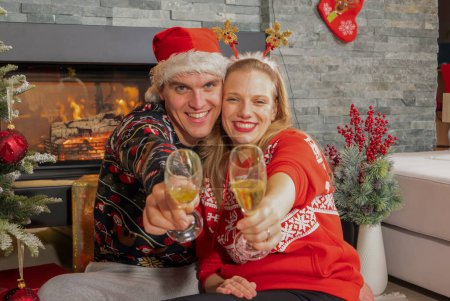 Téléchargez les photos : Lovely young couple toasting with glasses of champagne towards camera. Happy young man and woman enjoying and celebrating with champagne sitting by Christmas tree in front of a fireplace. - en image libre de droit