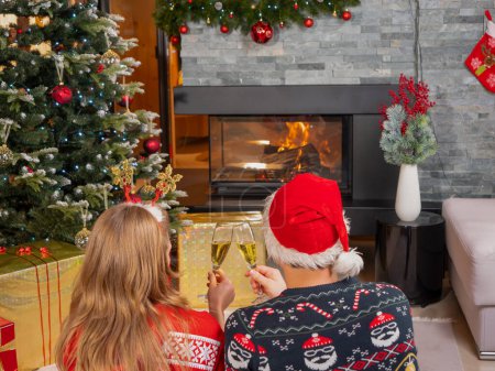 Téléchargez les photos : Couple in Christmas sweaters watching fireplace and toasting with champagne. Cheerful man and woman celebrating Christmas Eve in front of the fireplace in a beautifully decorated home living room - en image libre de droit