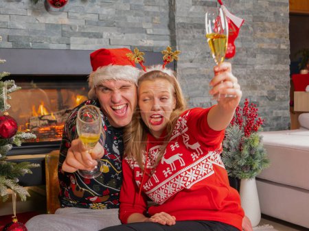Photo for Young couple makes silly faces and toasts with glasses of champagne. Cheerful man and woman enjoying and celebrating with champagne sitting by the Christmas tree in front of a fireplace. - Royalty Free Image