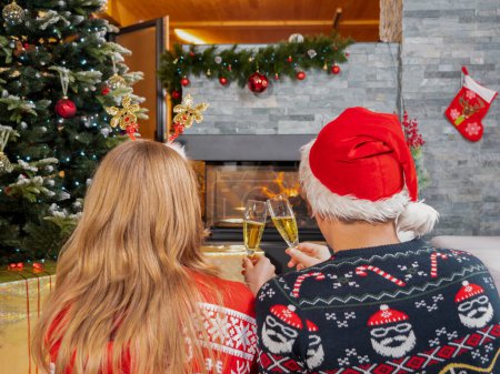 Foto de Young romantic couple looking at fireplace and toasting with champagne. Cheerful man and woman celebrating Christmas Eve in front of the fireplace in a beautifully decorated home living room - Imagen libre de derechos