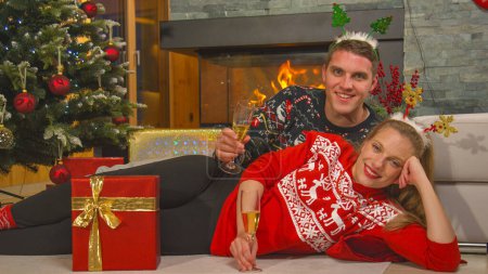Téléchargez les photos : Couple in festive mood with glasses of champagne lies among gifts. Merry young man and woman enjoying and celebrating with champagne while lying by Christmas tree in front of a fireplace. - en image libre de droit