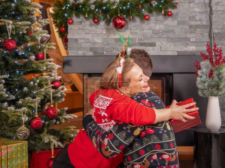 Foto de Grateful woman embraces her husband who made her happy with a present. Cheerful couple enjoy and celebrate Christmas holiday by the fireplace in a beautifully decorated home living room. - Imagen libre de derechos