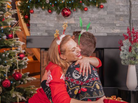 Foto de Beautiful woman hugs her husband who surprised her with Christmas gist. Cheerful couple celebrates winter holidays and exchanges presents on festive Evening in a beautifully decorated home. - Imagen libre de derechos