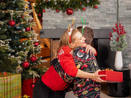 Foto de Happy woman in arms of a man who surprised her with Christmas gift. Cheerful couple enjoy and celebrate Christmas holiday by the fireplace in a beautifully decorated home living room. - Imagen libre de derechos