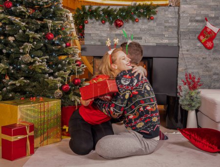 Téléchargez les photos : Pretty young lady hugging a man who surprised and delighted her with a gift. Lovely couple celebrate Christmas holiday and exchange presents by the beautifully decorated tree in front of fireplace. - en image libre de droit
