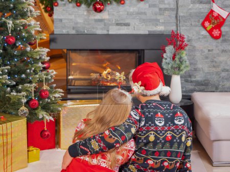 Téléchargez les photos : Rear view of romantic couple hugging and looking at fireplace on Christmas Eve. Young man and woman embrace and enjoy romance in front of the fireplace in beautifully decorated room on festive Evening - en image libre de droit