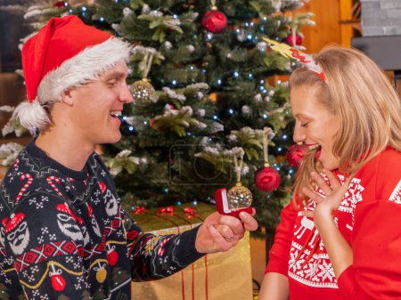 Téléchargez les photos : Young guy surprises his girlfriend with engagement ring on Christmas. Couple in love gets engaged under Christmas tree. Romantic gesture on festive Evening and cause for another celebration. - en image libre de droit