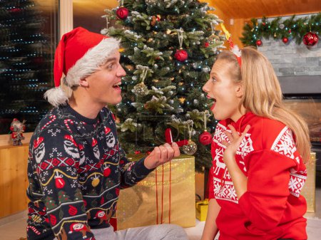 Téléchargez les photos : Loving couple on a romantic engagement under the Christmas tree. Young man surprises his lady with a wonderful romantic gesture and proposes to her with an engagement ring on Christmas. - en image libre de droit
