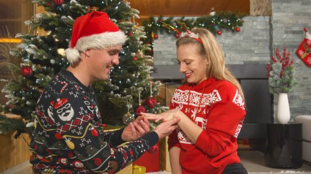 Téléchargez les photos : Happy and handsome man giving his beautiful lady an engagement ring on Christmas. Young man surprises his lady with wonderful romantic gesture and proposes to her with an engagement ring on Christmas. - en image libre de droit