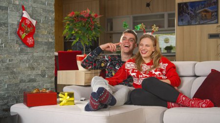 Téléchargez les photos : Laughing young couple sitting on comfy couch and watching Christmas comedy on TV. Man and woman relaxing on cosy sofa in home living room and enjoying at funny holiday television entertainment program - en image libre de droit
