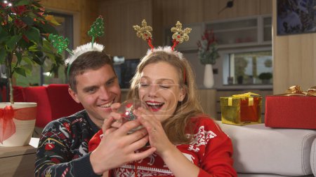 Téléchargez les photos : Cheerful man surprises his beautiful lady with a Christmas snow globe. Young couple in festive mood celebrating Christmas holidays and enjoying relaxing time together in home living room. - en image libre de droit