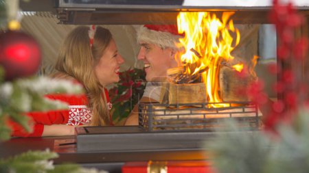 Foto de Cute couple in love showing affection while sitting by the burning fireplace. Cheerful man and woman wearing Christmas sweaters and enjoying romantic ambiance in home living room on Christmas Eve. - Imagen libre de derechos
