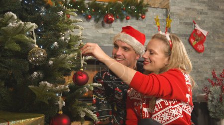 Photo for Smiling romantic couple decorating Christmas tree with red baubles at fireplace. Young man and woman preparing Christmas decoration in their home living room for celebrating festive winter holidays. - Royalty Free Image