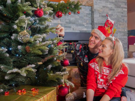 Photo for Romantic couple sitting at fireplace and decorating Christmas tree with baubles. Young man and woman preparing Christmas decoration in their home living room for celebrating festive winter holidays. - Royalty Free Image