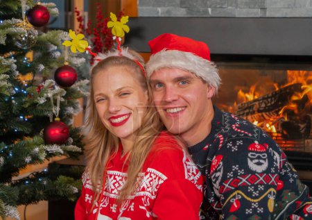 Téléchargez les photos : Joyful festive couple posing in front of fireplace by Christmas tree. Handsome man and beautiful woman in pose, wearing Christmas sweaters and smiling into camera for December postcard. - en image libre de droit