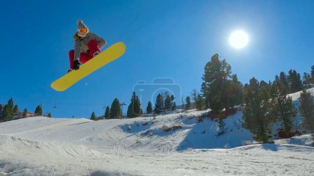 Foto de Freestyle female snowboarder performing stylish indy grab trick. Young woman with snowboard jumping on kicker at snow park in mountain ski resort. Sunny winter day for snowboarding. - Imagen libre de derechos
